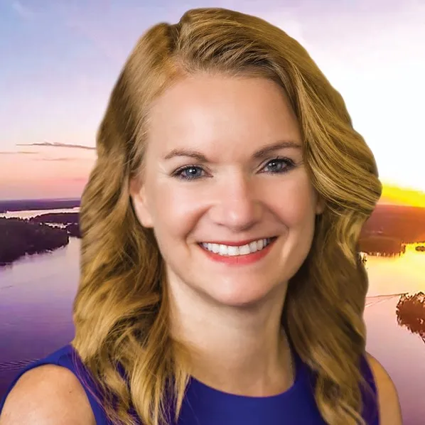 Smiling woman with blonde hair and blue eyes in front of a sunset over a river landscape
