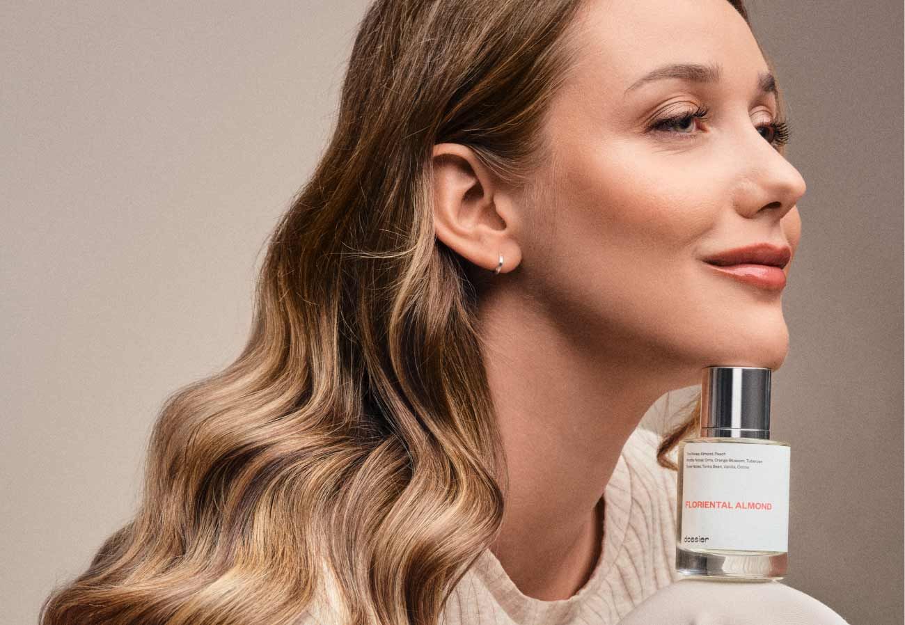Woman with wavy blonde hair holding a bottle of Floriental Almond perfume against a neutral background