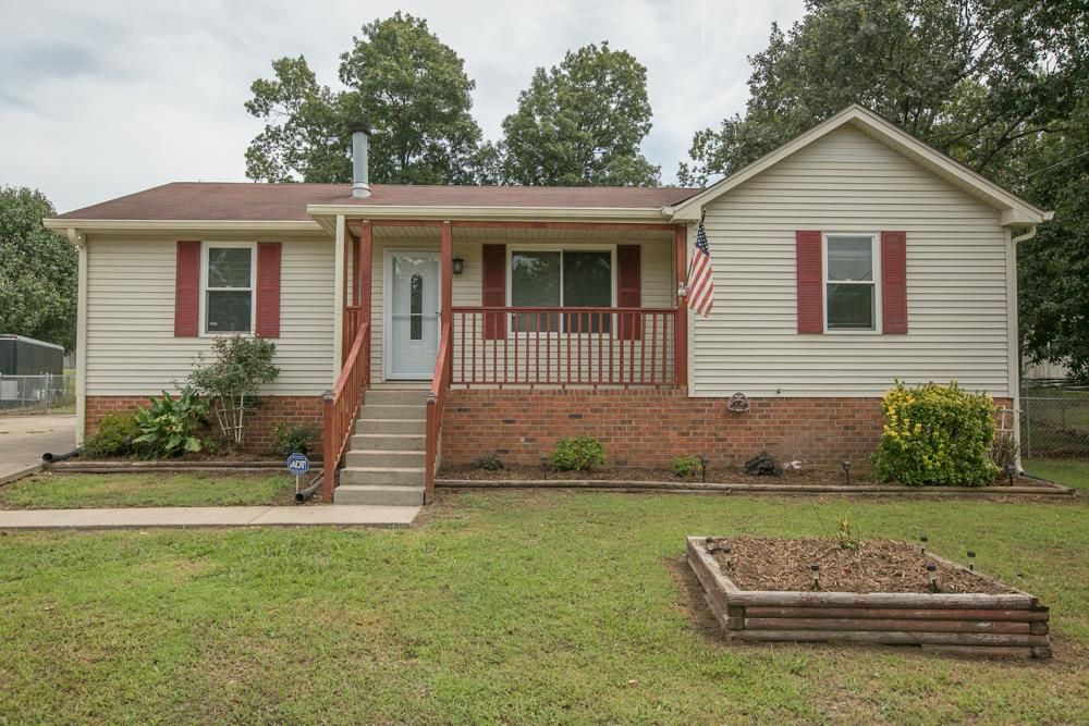 Single-story house with beige vinyl siding, red shutters, a front porch with stairs, and an American flag, surrounded by a green lawn and trees in the background.