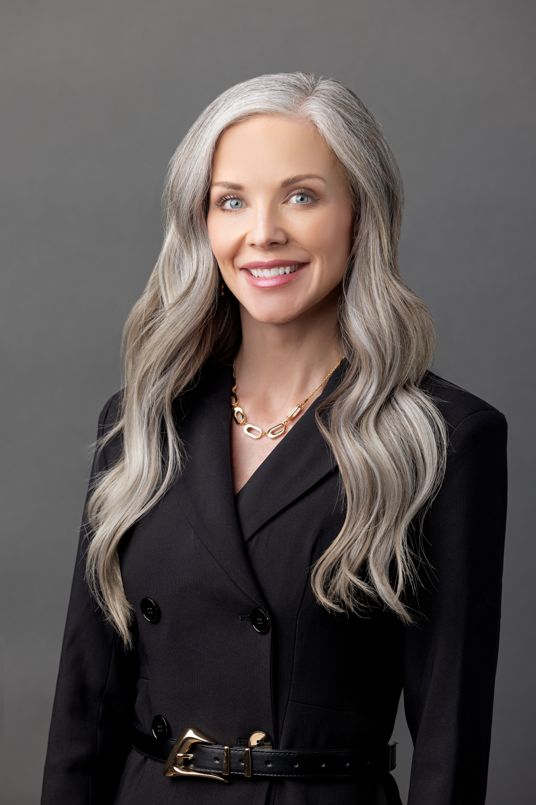 Smiling woman with long blonde hair wearing a sleeveless light blue dress against a plain light gray background