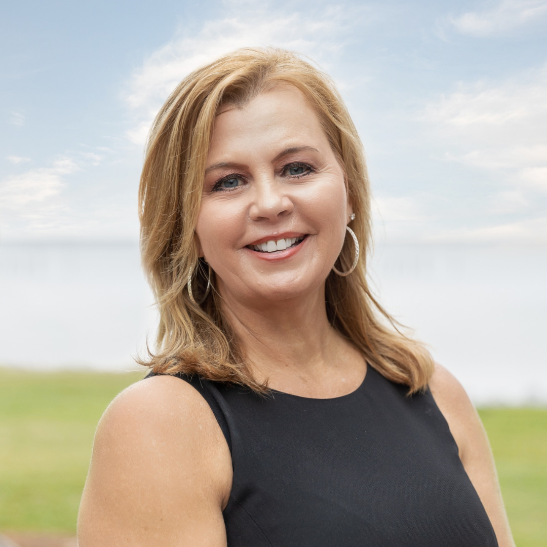 Smiling woman with shoulder-length red hair wearing a black blazer and white top, outdoors on a sunny day.