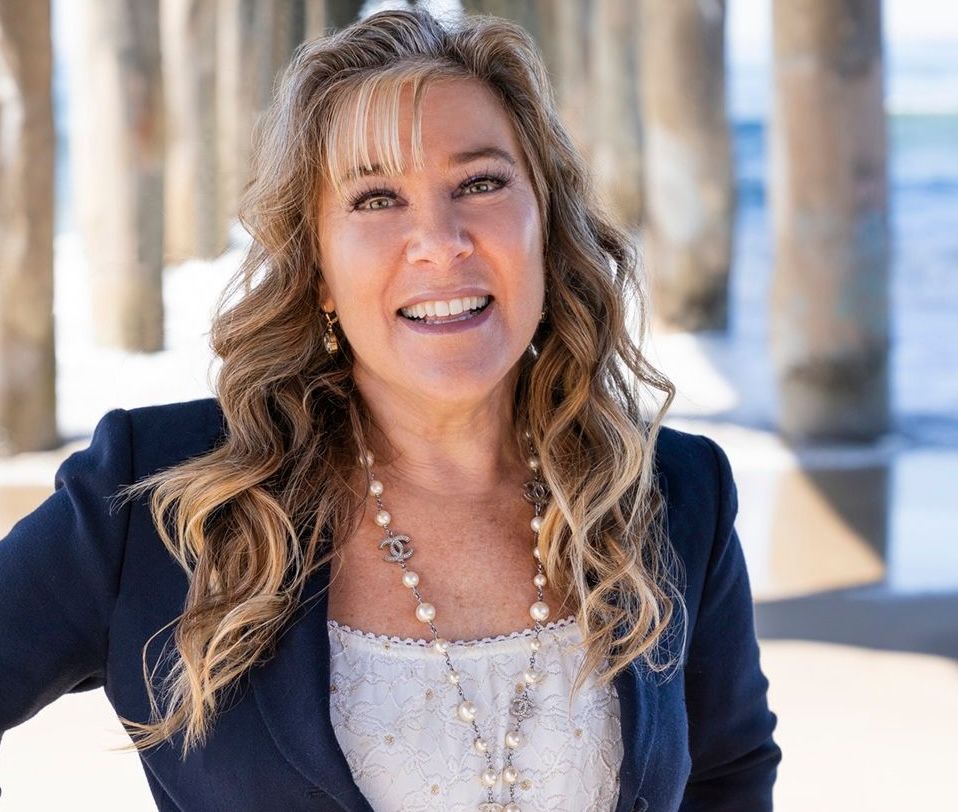 Portrait of a professional woman with blonde curly hair, wearing a dark blazer and pearl jewelry, standing in front of large green tropical leaves.
