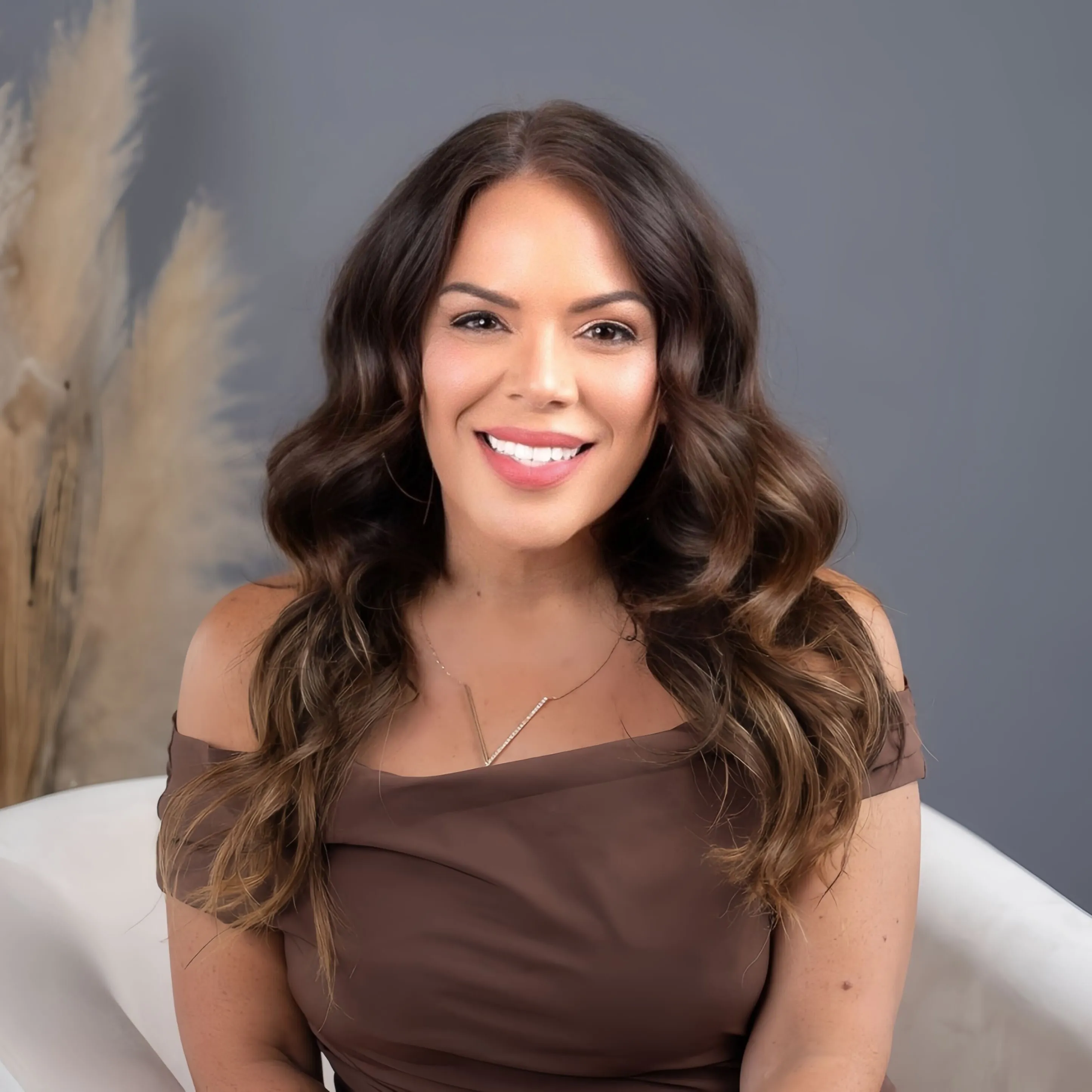 Portrait of a young woman with wavy brown hair, smiling softly, wearing a brown off-shoulder top and a delicate gold necklace against a dark grey background.