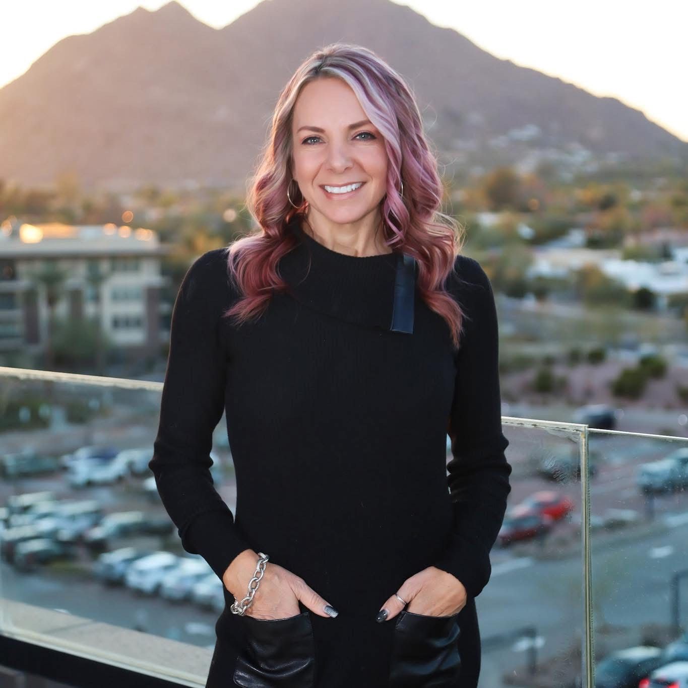 Smiling woman with long wavy hair in pink and blonde shades wearing a black long-sleeve top posing against a city background in the sunset.