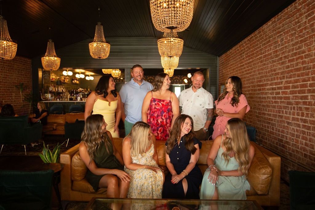 Group of professional women and one man posing together in a modern kitchen, dressed in business casual attire, smiling at the camera.