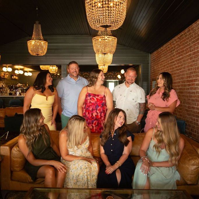 Group of professional women and one man posing together in a modern kitchen, dressed in business casual attire, smiling at the camera.