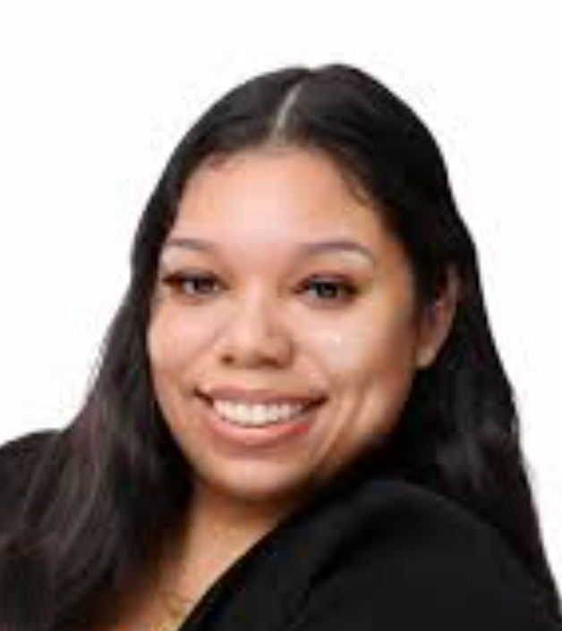 Smiling woman with long dark red hair wearing large beaded hoop earrings, a gray jacket, and layered necklaces outdoors with greenery in the background.