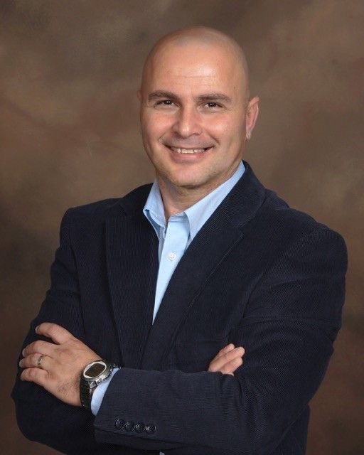 Professional businessman in a dark suit and light blue shirt smiling with arms crossed against a brown background