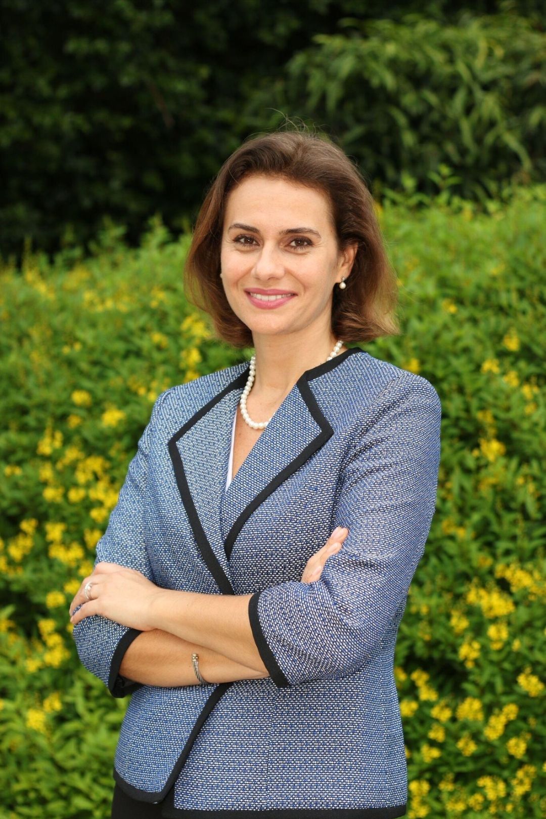 Professional woman smiling outdoors in a blue blazer with a pearl necklace, standing in front of green foliage and yellow flowers.