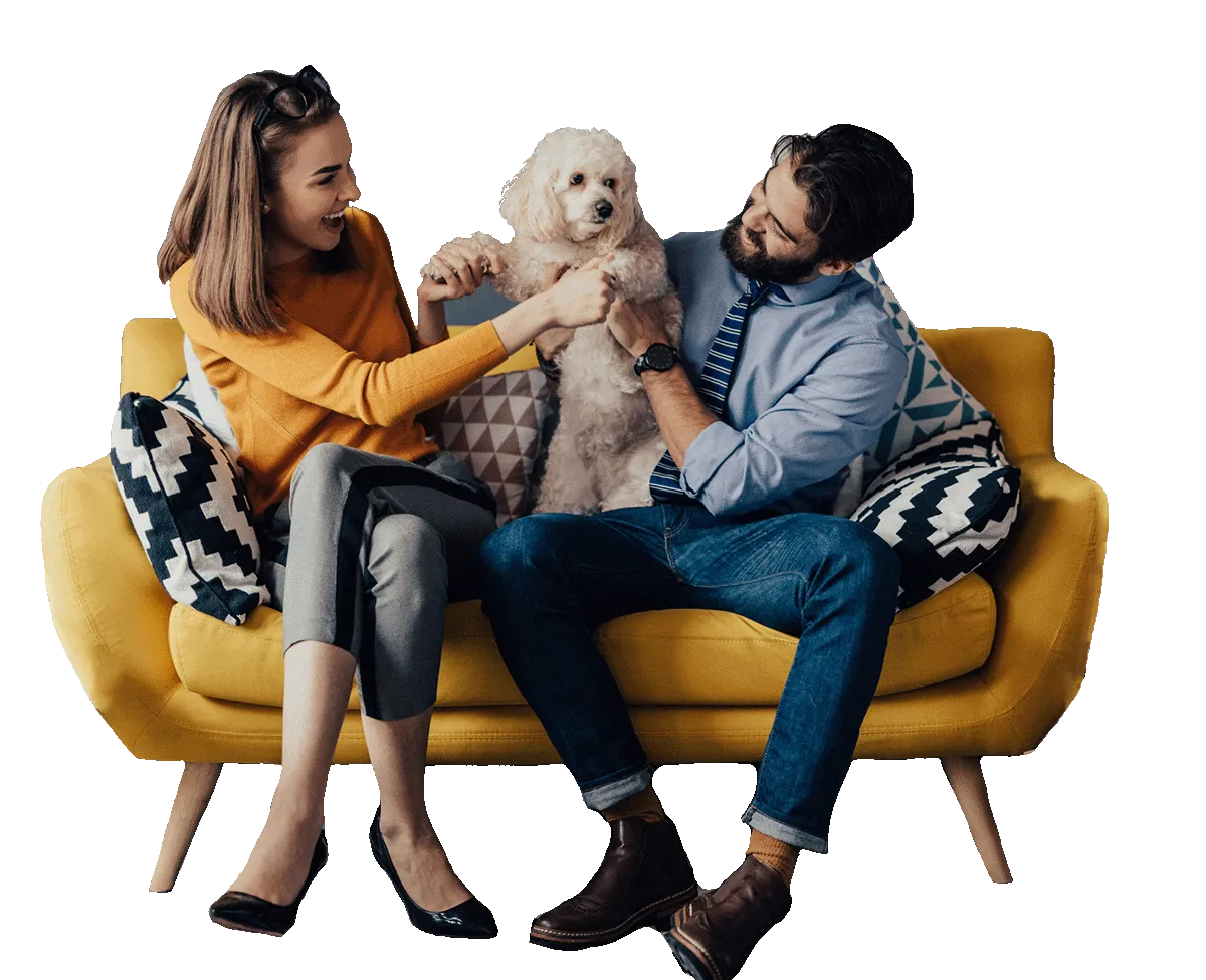 Happy couple sitting on a yellow couch playing with a small, fluffy, cream-colored dog.