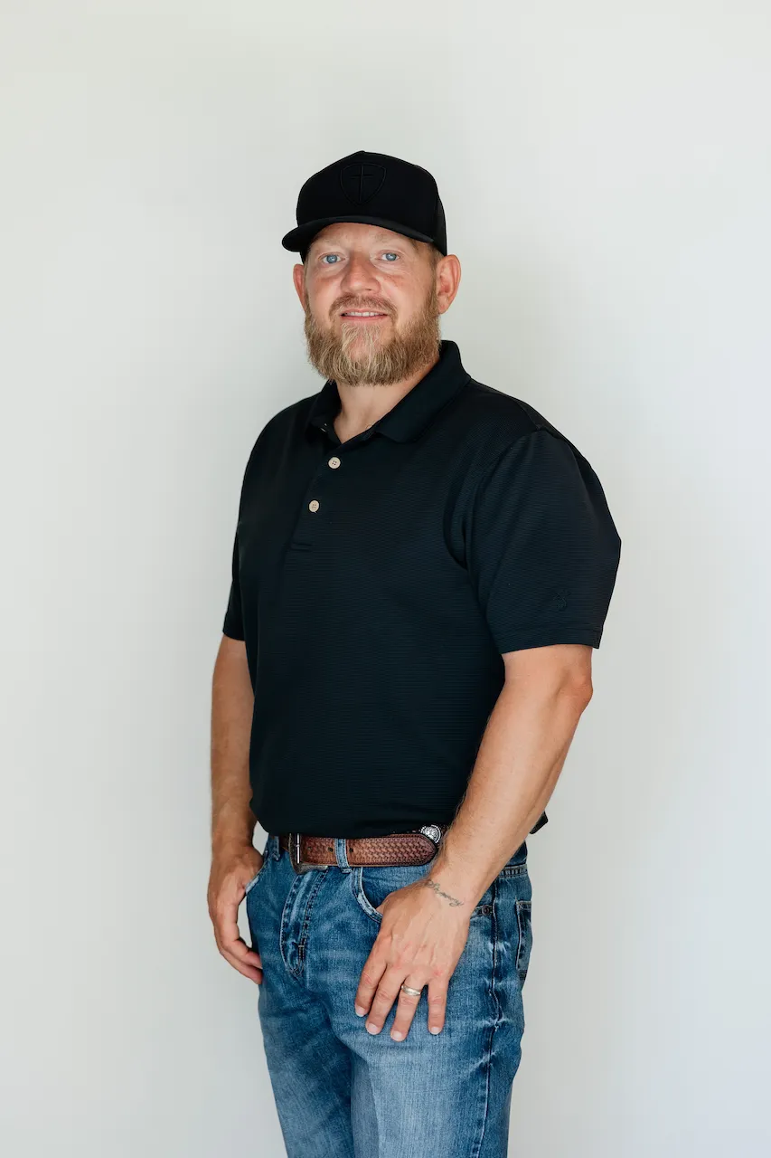 Portrait of a man in a black cowboy hat with a beard, wearing a black vest over a patterned shirt, standing against a light gray background