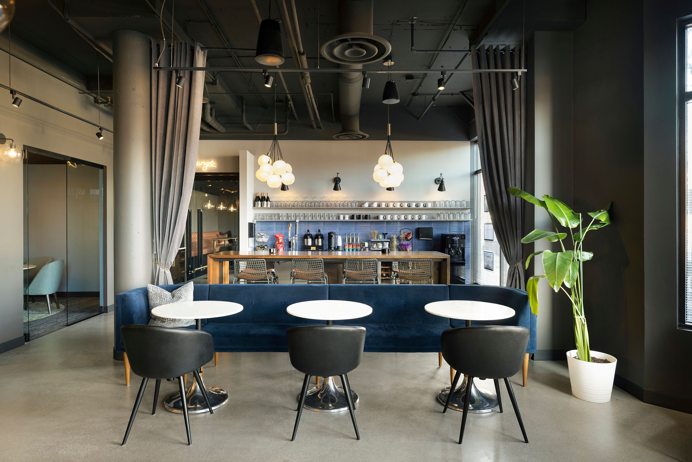 VIBE offices. Three white tables with black chairs and a blue couch in foreground. Bar with drinks, snacks, mugs and glasses in background.