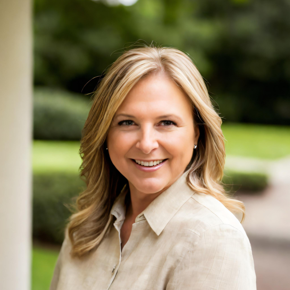 Portrait of a smiling middle-aged woman with wavy blonde hair wearing a navy blue blouse with blue and white floral patterns and a pearl necklace outdoors in a park setting.