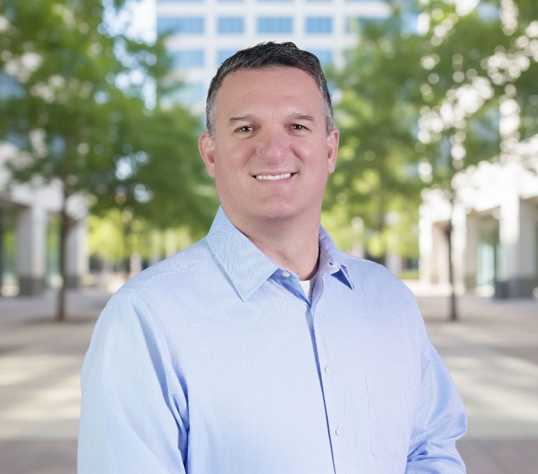 Professional portrait of a man in a gray suit and blue shirt with a striped tie, smiling against a light yellow background