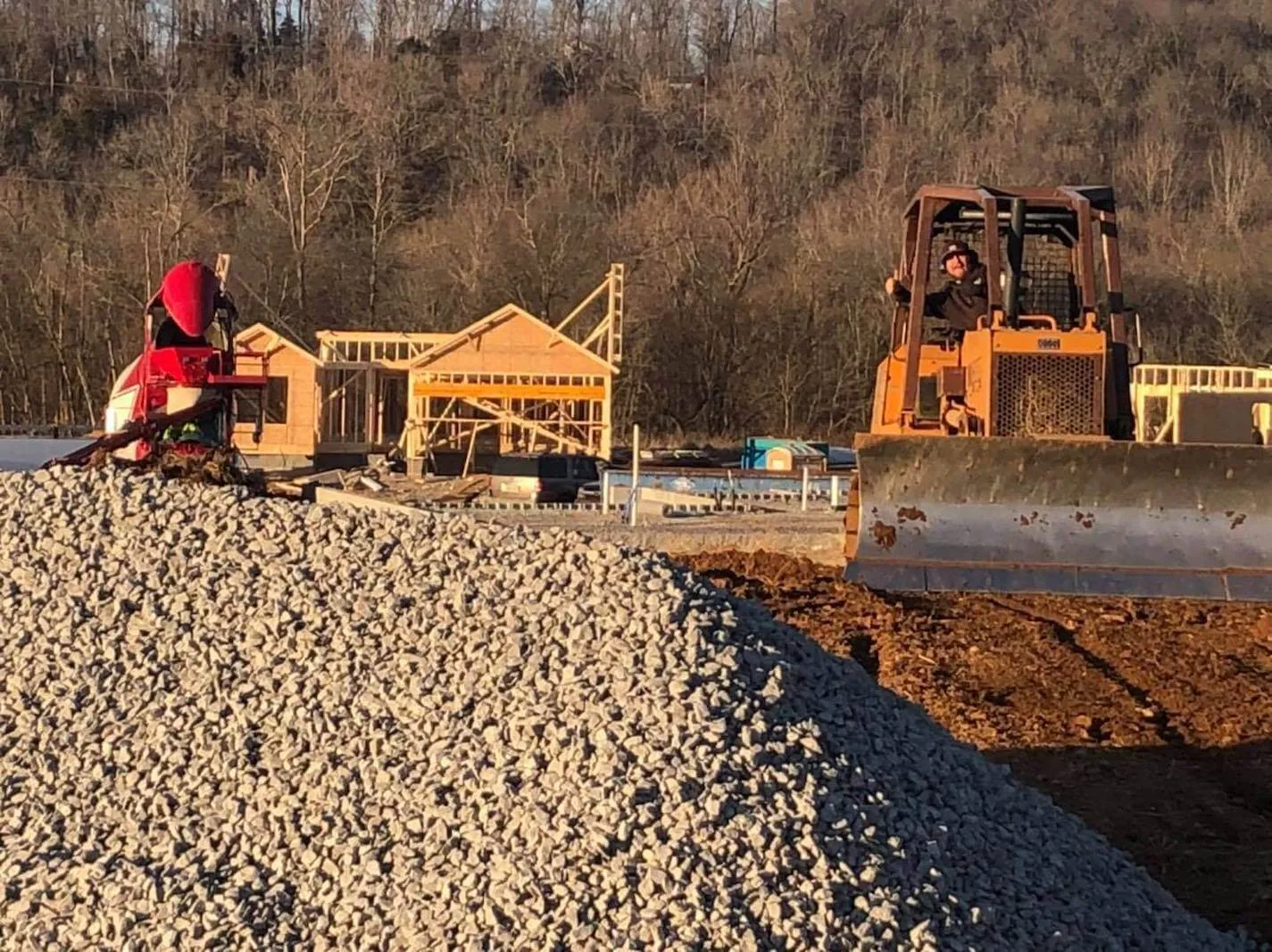 Construction site with a bulldozer, gravel pile, and partially built wooden structures in a rural area during late afternoon.