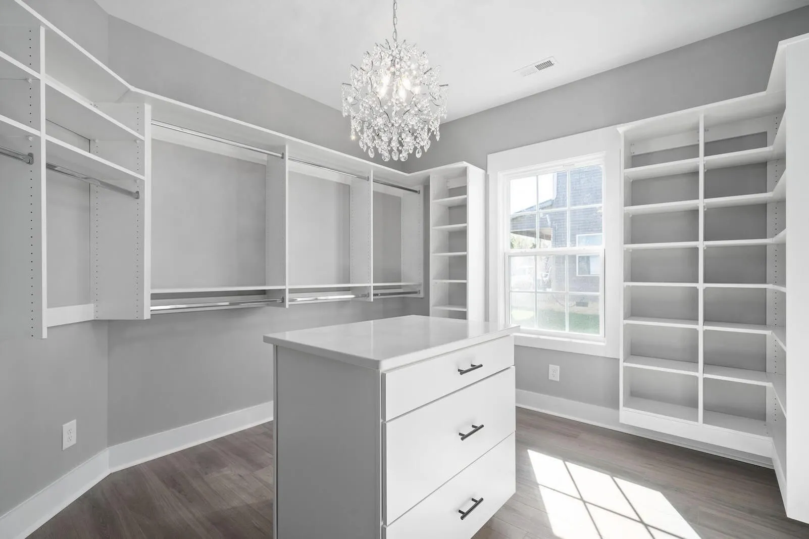 Spacious walk-in closet with white shelving, dresser, large window, and chandelier, featuring modern design and ample storage space.
