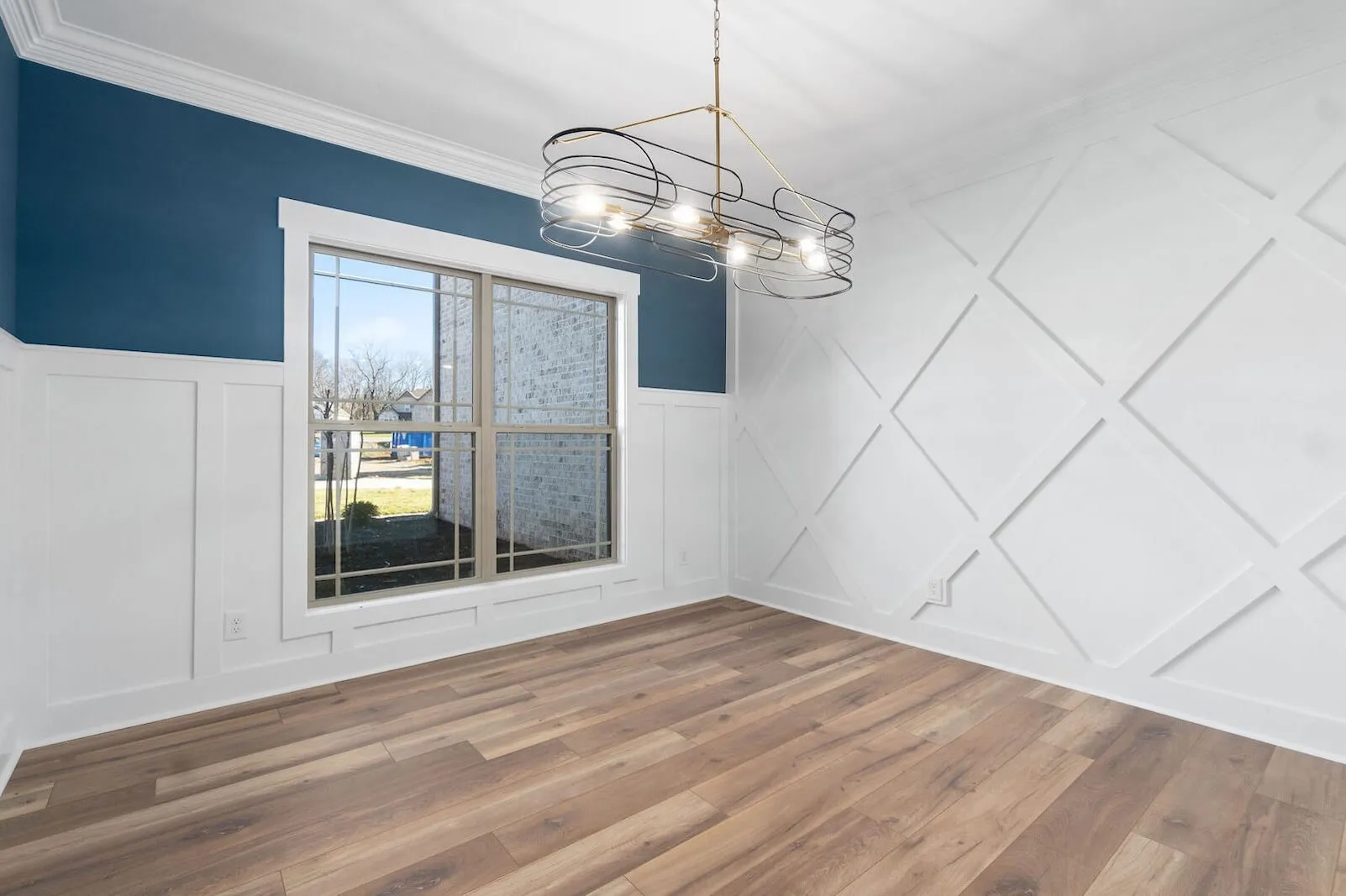 Bright empty dining room with large window, white paneled and blue accent walls, wood flooring, and modern chandelier.