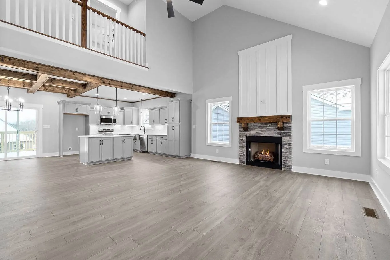 Open floor plan living room and kitchen with modern design, stone fireplace, wooden beams, large windows, and light gray walls.
