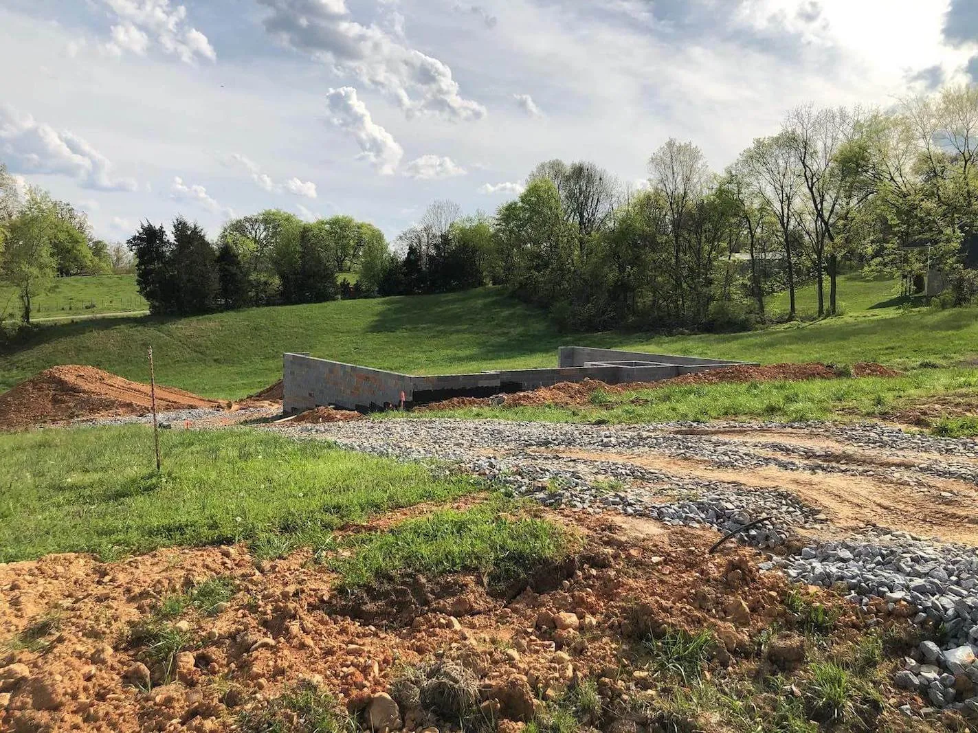 Construction site in a rural area with a partially built concrete structure, dirt, gravel, green grass, and trees under a partly cloudy sky.
