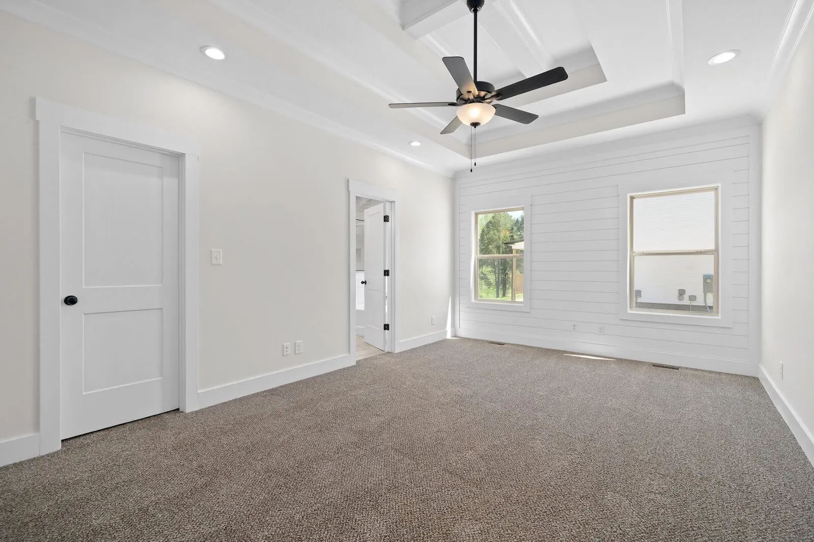 Empty modern living room with white walls, beige carpet, ceiling fan, recessed lighting, and two large windows overlooking a green yard