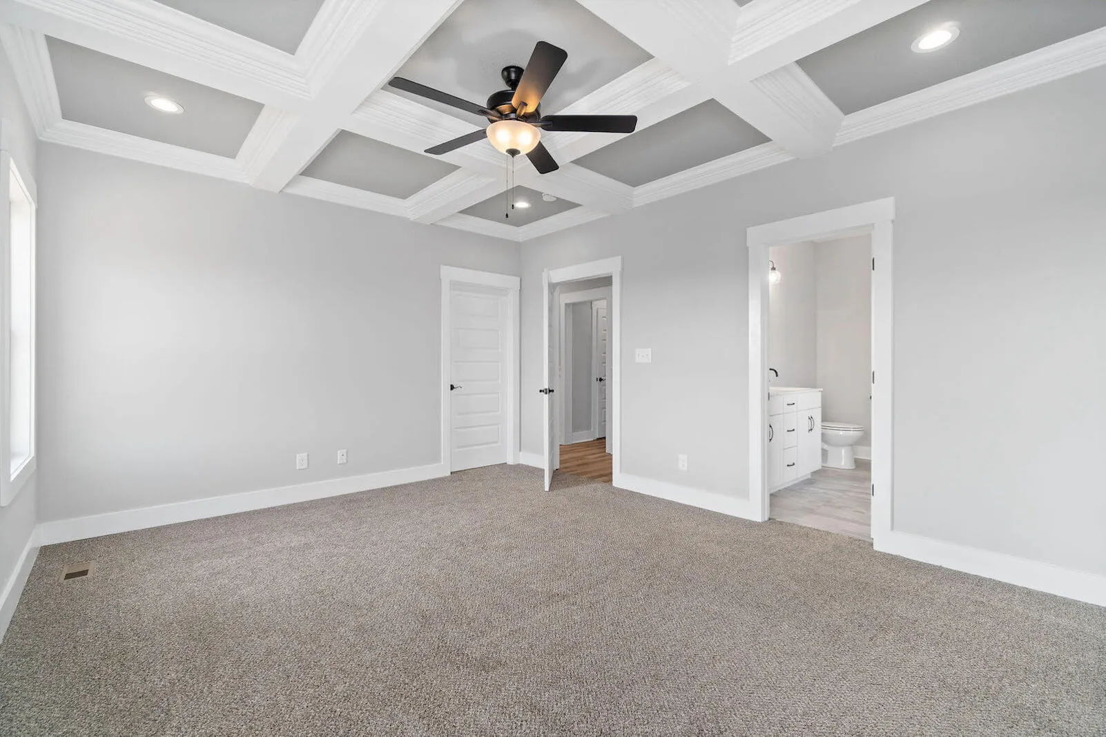 Spacious modern bedroom with neutral-colored carpet, coffered ceiling with ceiling fan, and attached bathroom with vanity and toilet