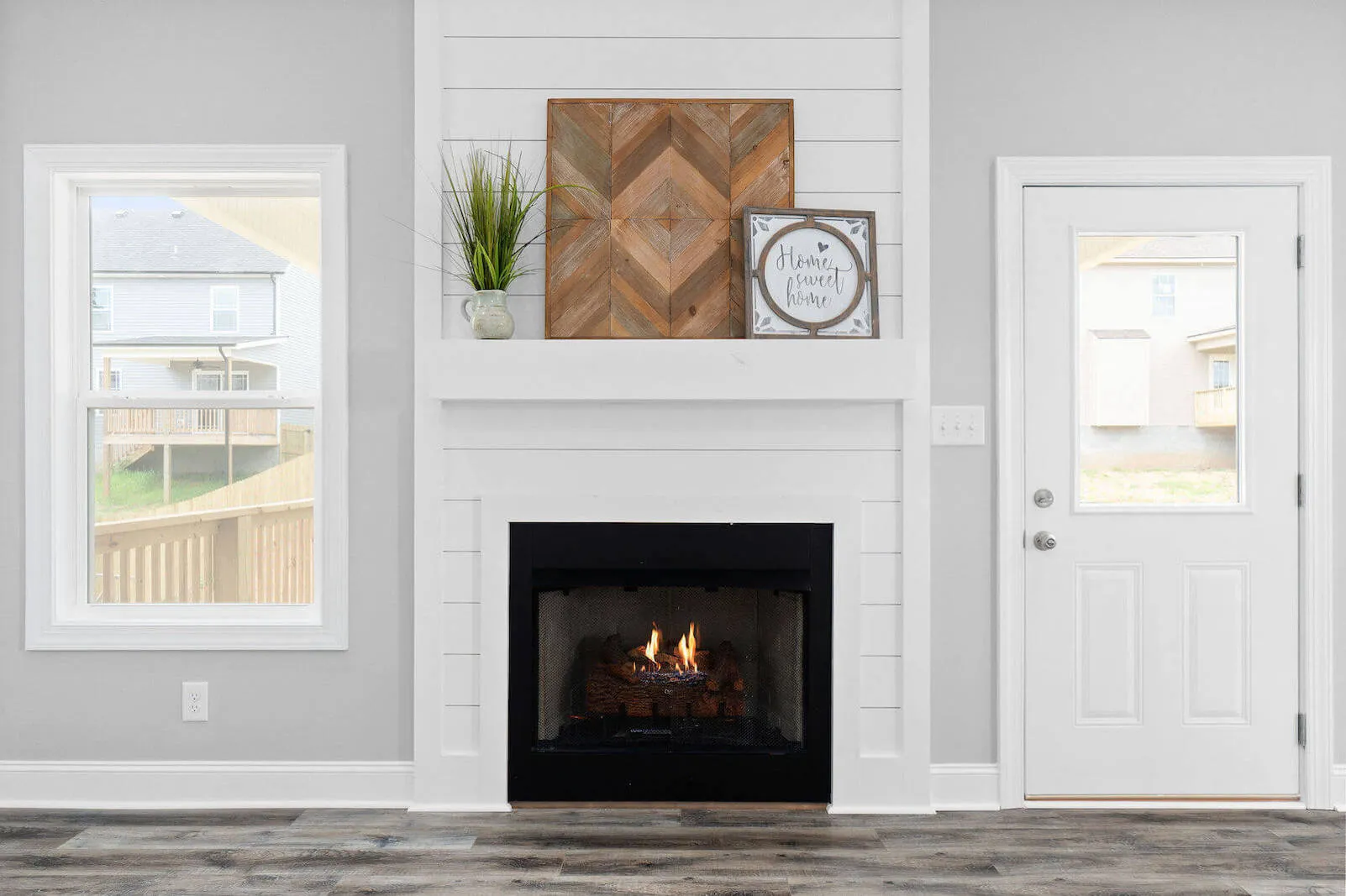 Modern living room interior with a white fireplace, wooden decorations, potted plant, and windows showing neighboring houses.