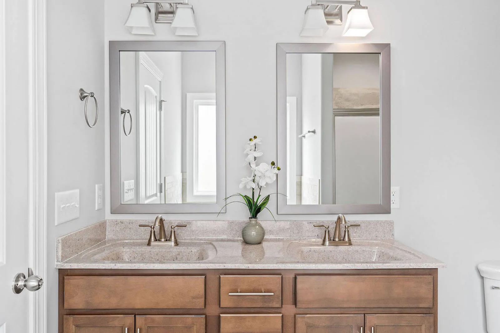 Double vanity bathroom with two mirrors, modern light fixtures, a beige granite countertop, wooden cabinets, and a decorative white orchid plant between the mirrors.