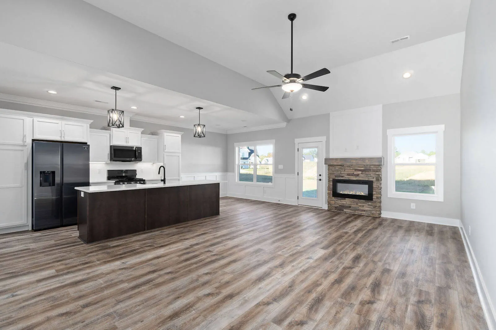 Spacious open-concept living room and kitchen with white cabinetry, dark island, stainless steel appliances, stone fireplace, and hardwood flooring.