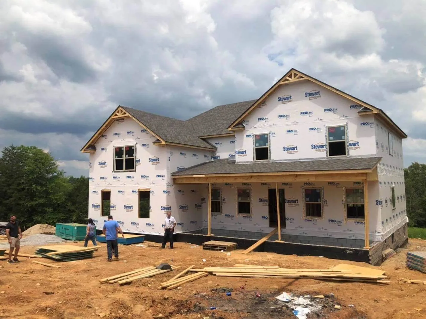 New construction of a two-story house with a partially finished exterior, showing windows and roof, on a dirt lot with construction workers and building materials under a cloudy sky.