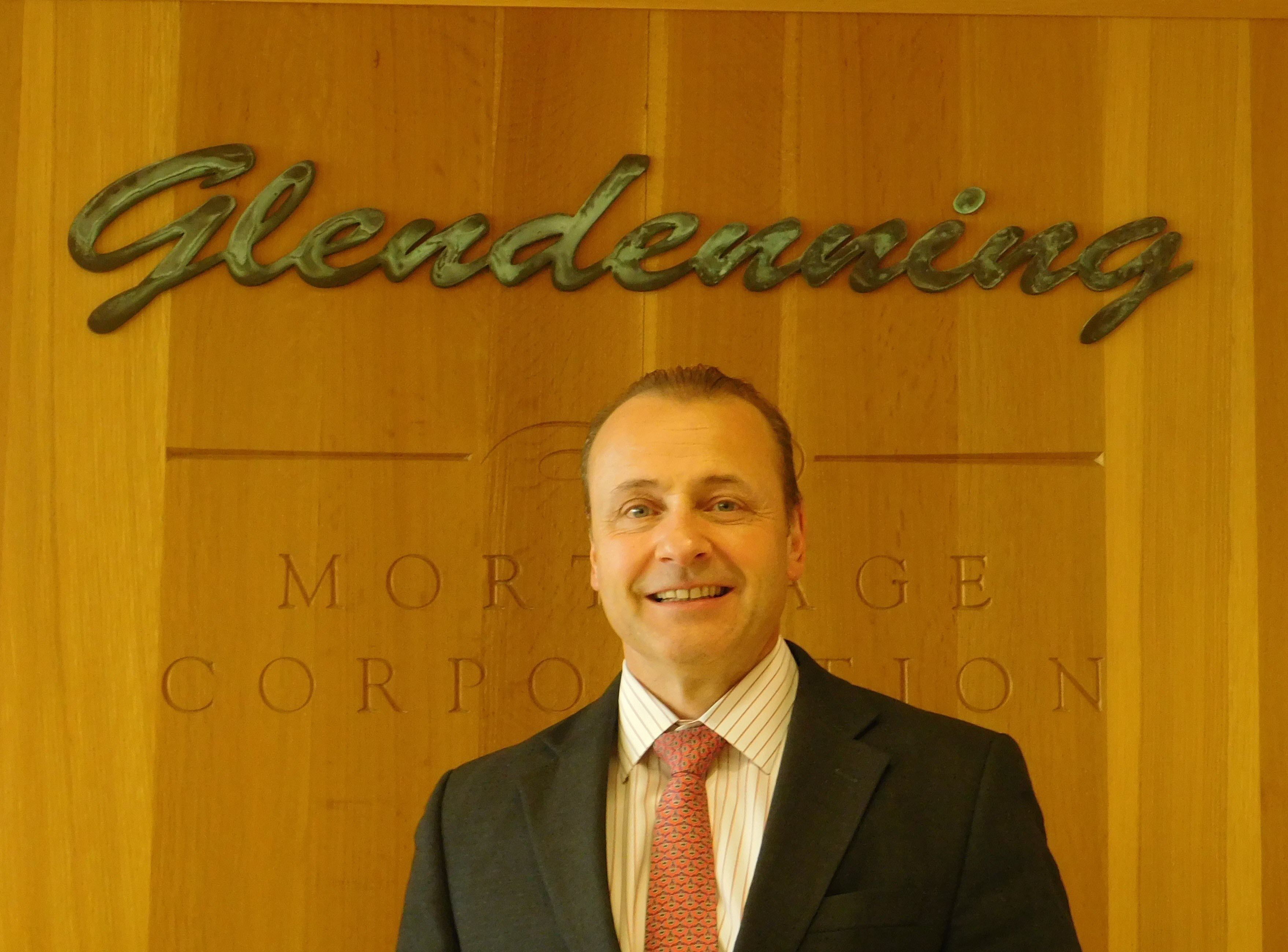 Smiling man in formal business attire standing in front of a wooden wall with the words 