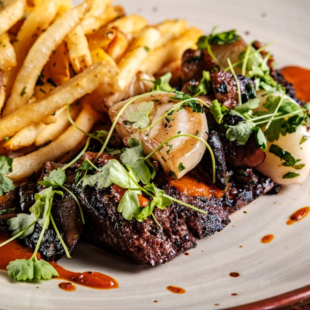 Plate with hamburger, French fries, lettuce, tomato, and onion sandwich with side of French fries on a white table, accompanied by a glass of dark soda, ketchup, and salt and pepper shakers.