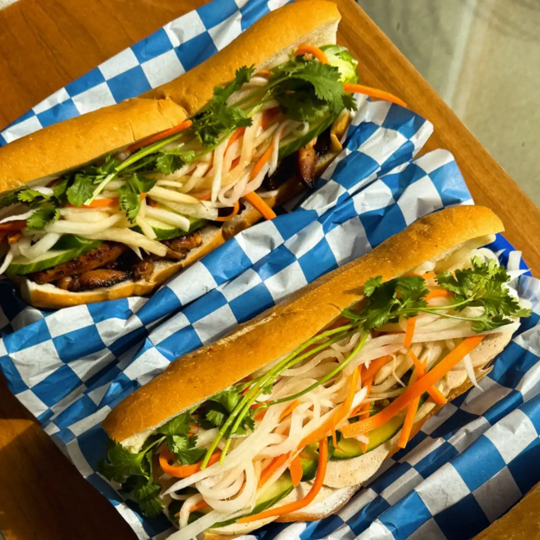 Two Vietnamese bánh mì sandwiches filled with grilled meat, fresh herbs, assorted pickled vegetables, and cucumber in crusty baguettes on blue-and-white checkered paper on a wooden surface.