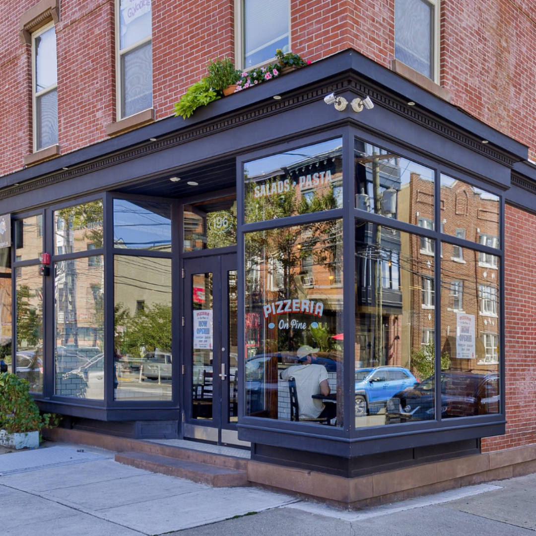 Exterior view of a corner pizzeria restaurant with large glass windows, brick upper walls, and potted plants on the window ledge, featuring signs for salads, pasta, and pizza.