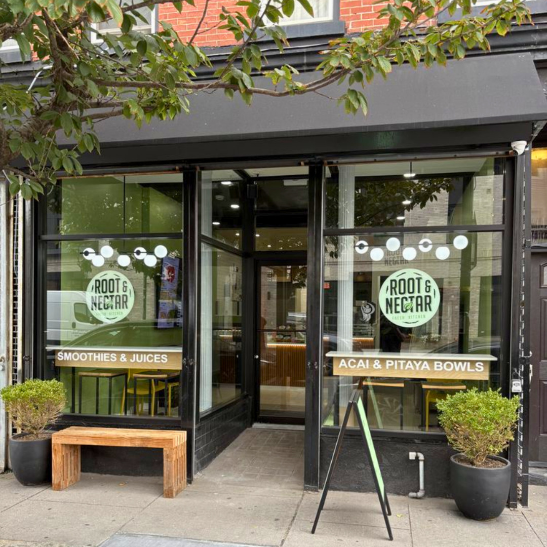 Exterior view of Root & Nectar juice bar and smoothie cafe with large glass windows, signage for smoothies, juices, acai, and pitaya bowls, potted plants, and a wooden bench outside.