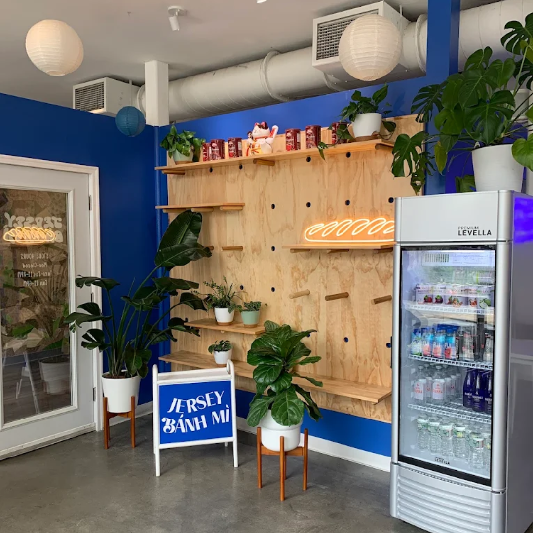 Modern indoor cafe corner with blue walls, wooden shelves, potted plants, a neon sign, and a glass-front refrigerator filled with beverages.