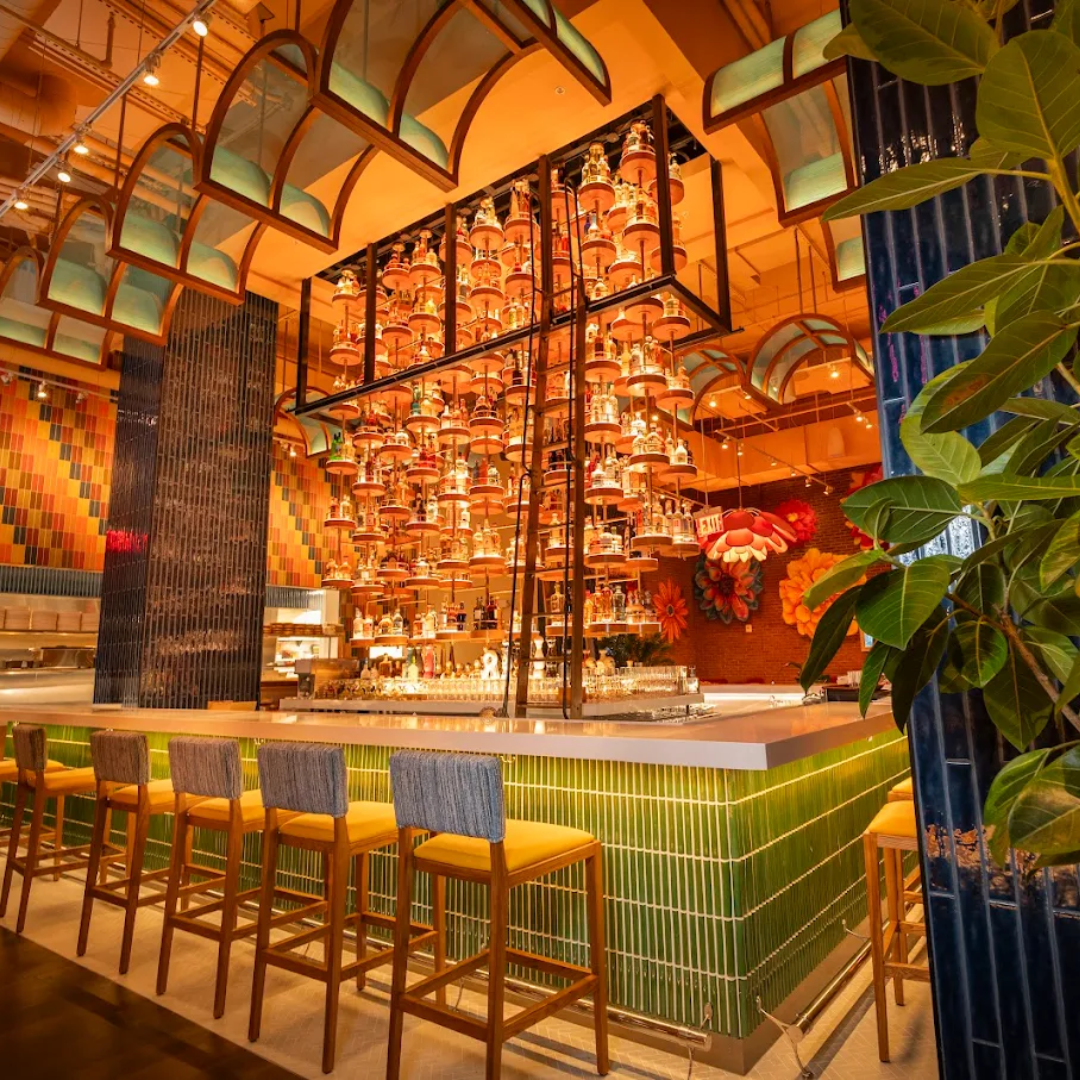 Colorful modern bar with a chandelier made of multiple small bottles, green tiled bar counter, wooden chairs with upholstered seats, and lush green plant in a vibrant, stylish restaurant interior.