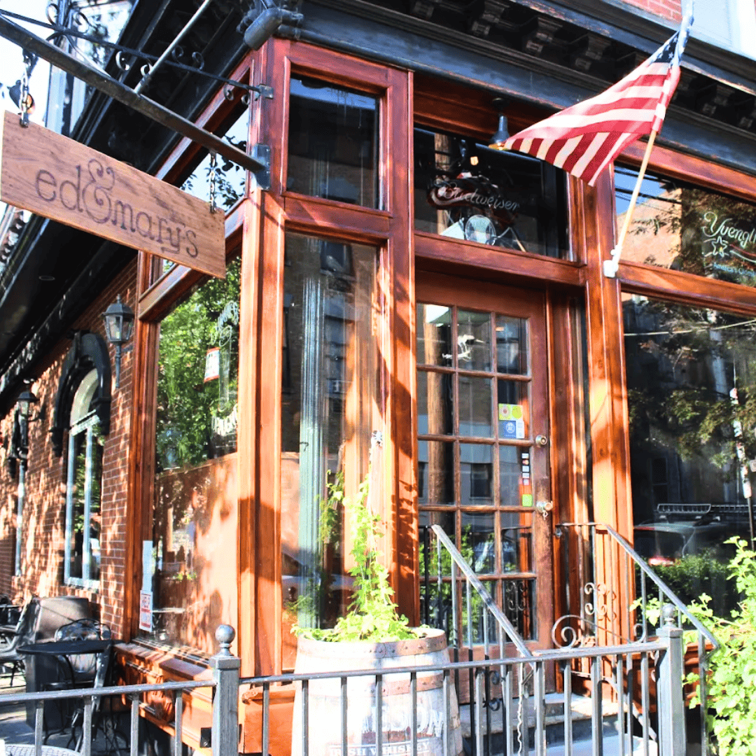 Exterior view of a corner pizzeria restaurant with large glass windows, brick upper walls, and potted plants on the window ledge, featuring signs for salads, pasta, and pizza.
