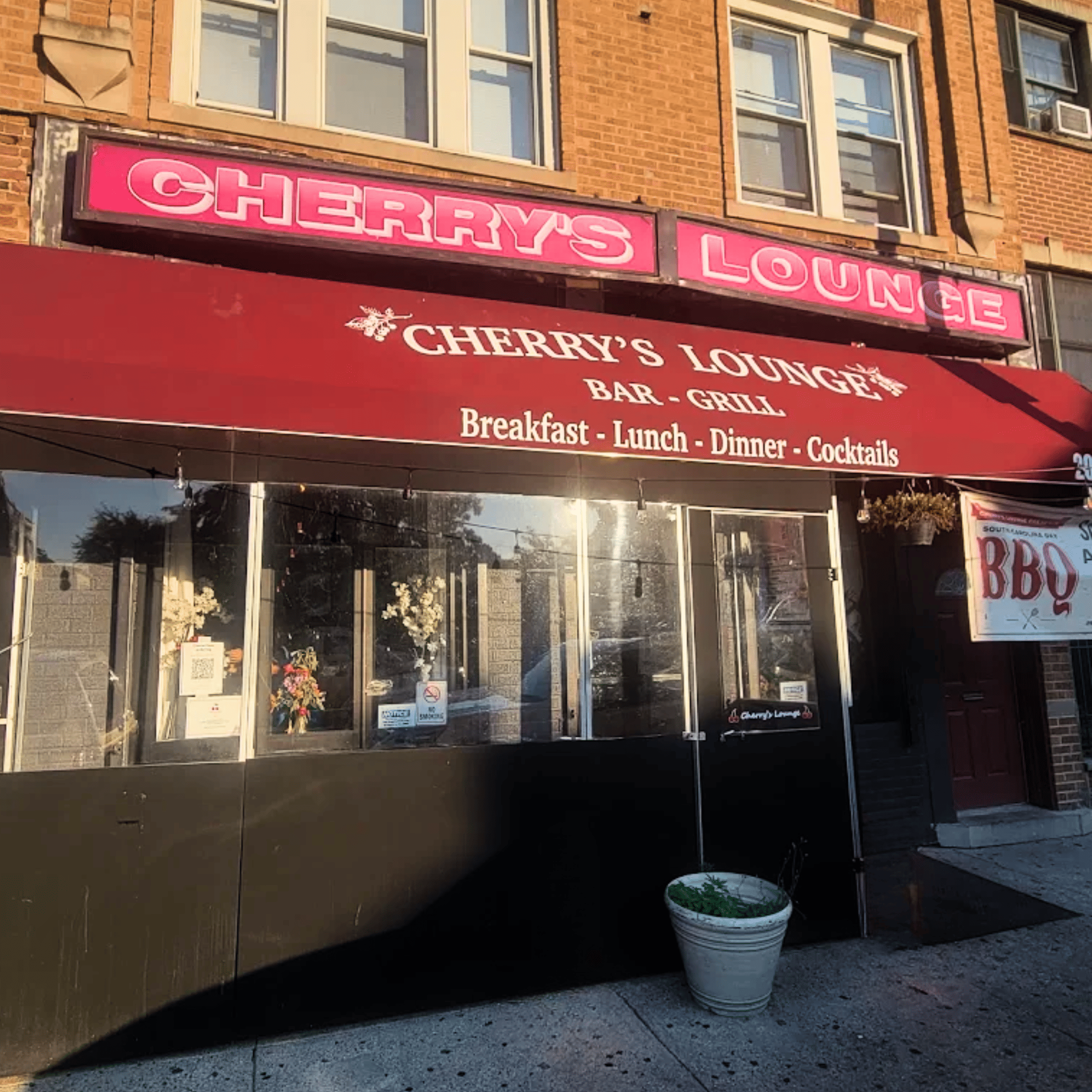 Exterior view of a corner pizzeria restaurant with large glass windows, brick upper walls, and potted plants on the window ledge, featuring signs for salads, pasta, and pizza.