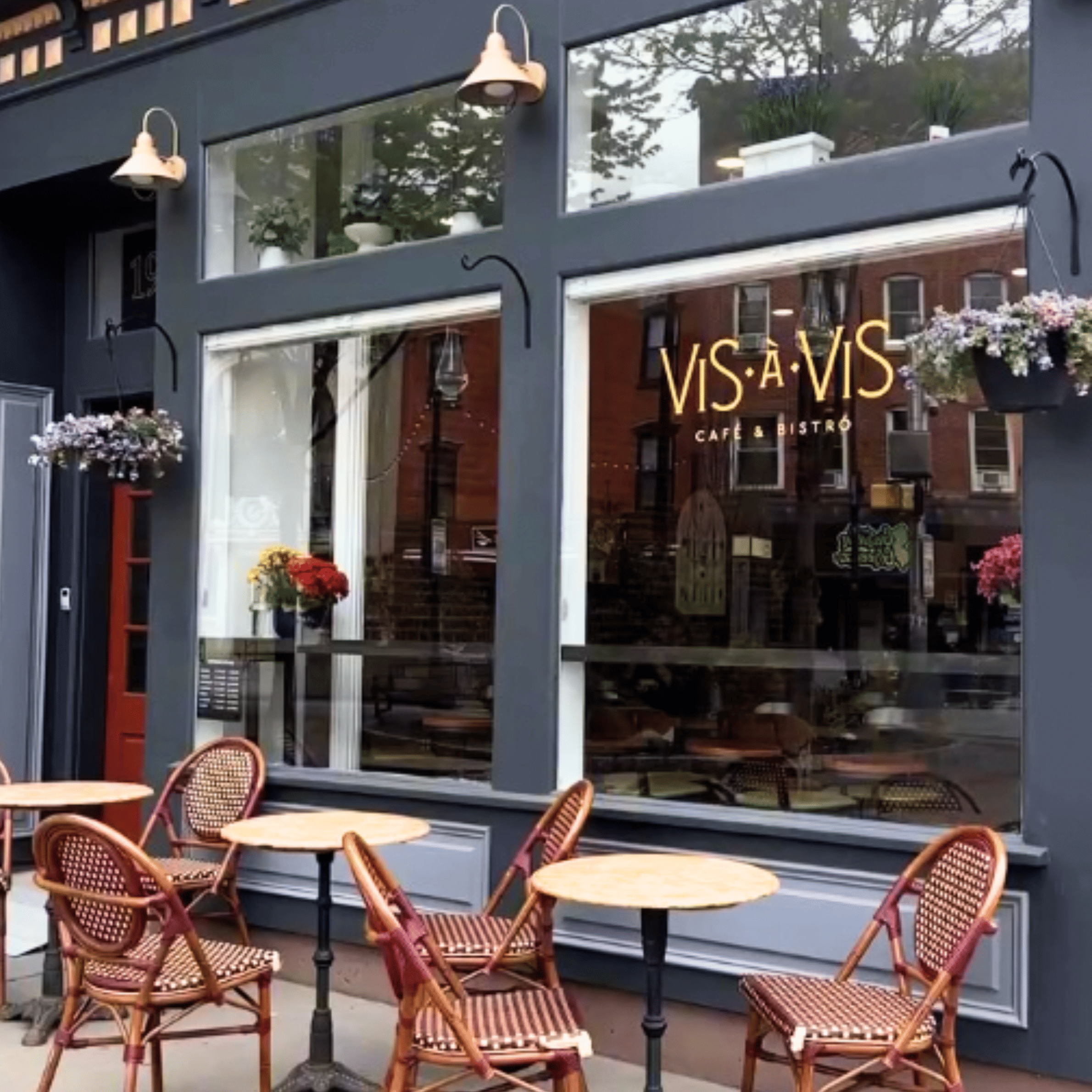 Exterior view of a corner pizzeria restaurant with large glass windows, brick upper walls, and potted plants on the window ledge, featuring signs for salads, pasta, and pizza.