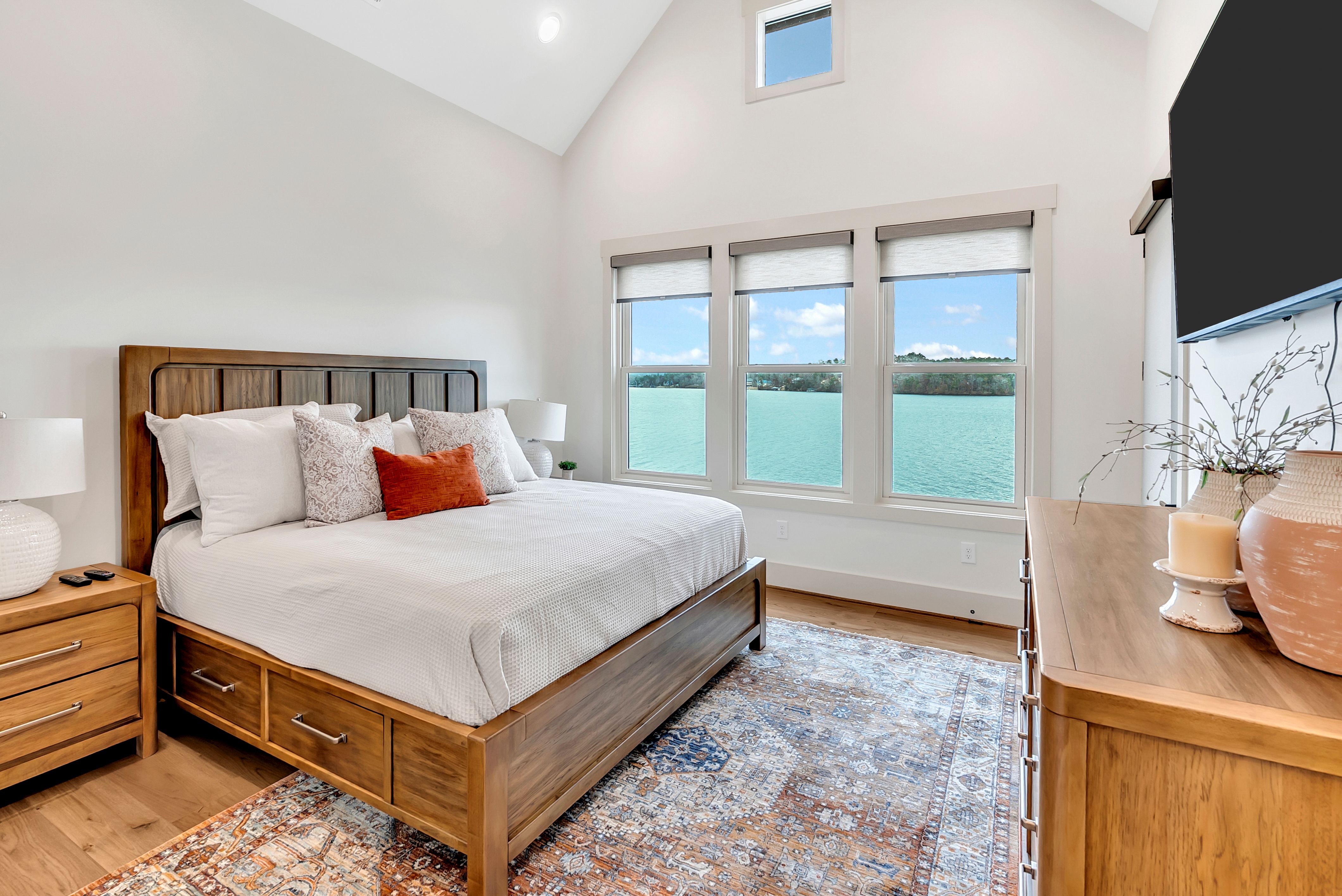Cozy bedroom with a large wooden bed, multiple pillows, and scenic lake view through three windows, decorated with neutral tones and modern furniture.