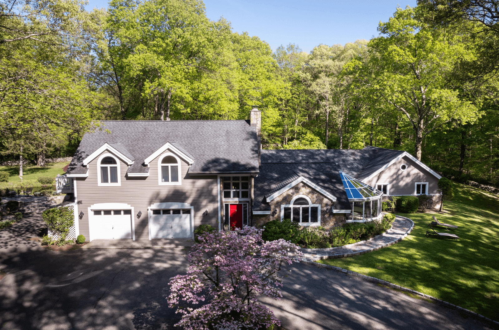 A spacious two-story house with gray siding, a red front door, a glass-enclosed sunroom, surrounded by lush green trees and a well-maintained lawn with pink flowering trees, under a clear blue sky.