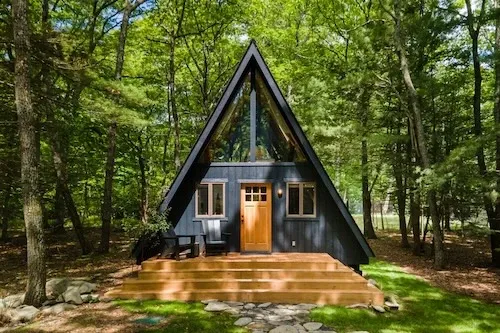A modern black A-frame tiny house with wooden accents and a front porch with steps, surrounded by lush green trees in a forest setting.