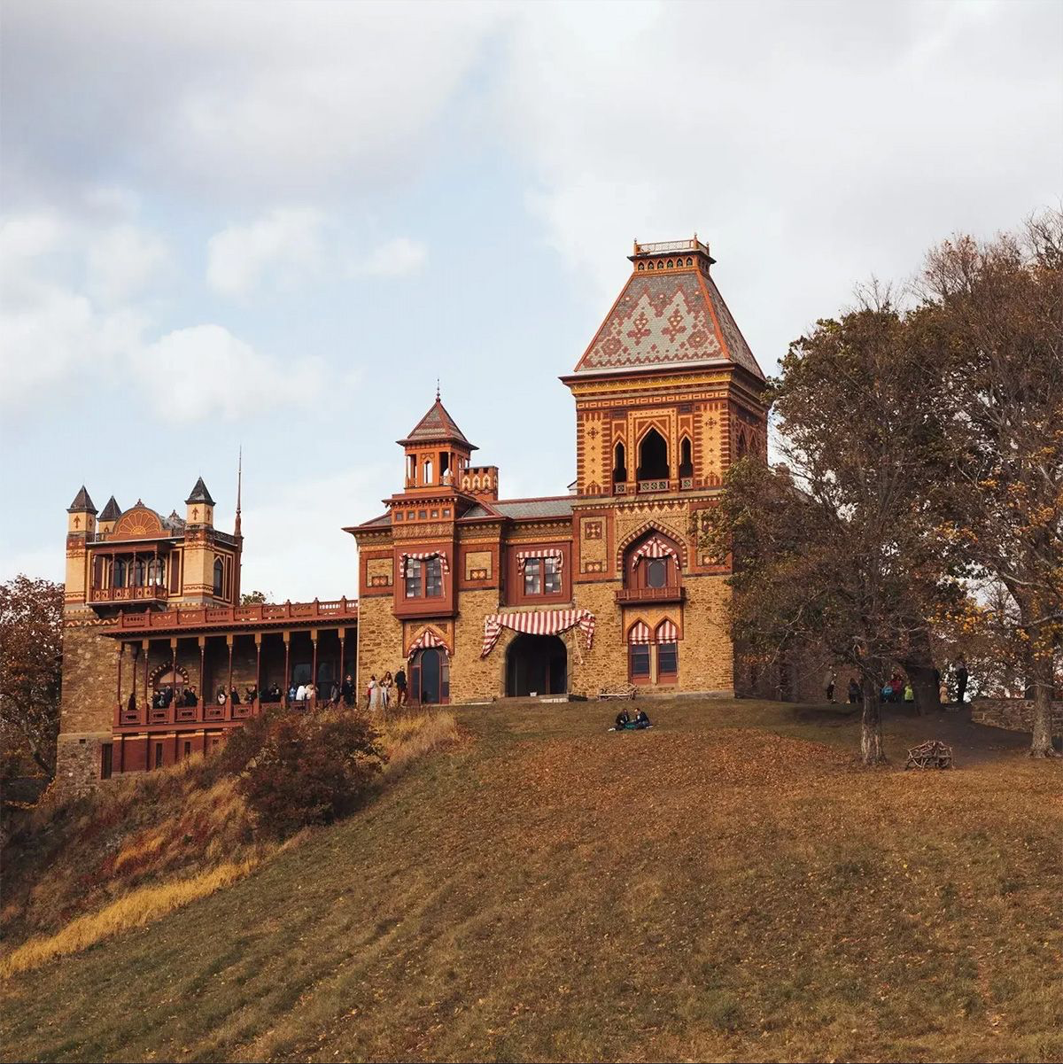 Fairy-tale castle with colorful brick architecture and turrets surrounded by autumn trees and a grassy hill in the background