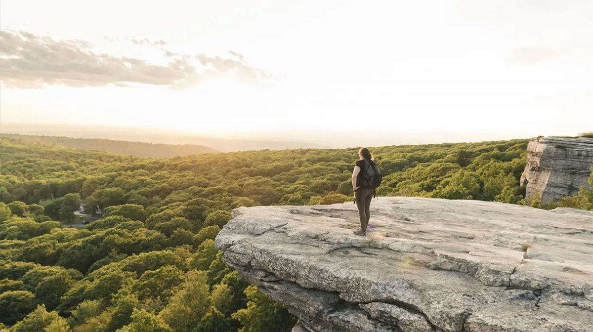 Hiker standing on a rocky cliff overlooking a lush green forest during sunset with a backpack and natural landscape.