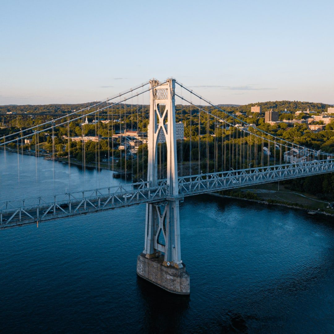 Aerial view of a large suspension bridge spanning over a river with a city skyline and green hills in the background.