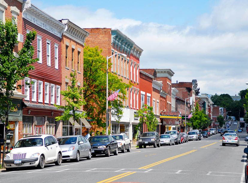 Historic downtown street with colorful brick buildings, parked cars, and green trees under a partly cloudy sky
