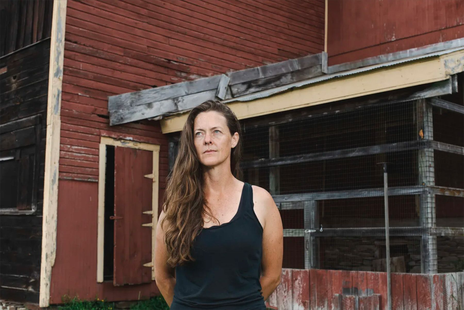 Portrait of a woman with long wavy brown hair wearing a black sleeveless top standing outdoors near a weathered red and black wooden barn with a chicken coop.