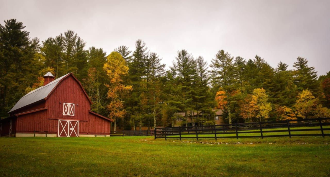Red wooden barn with white doors and trim on a grassy field surrounded by a black fence, set against a backdrop of green trees with autumn foliage under a cloudy sky.