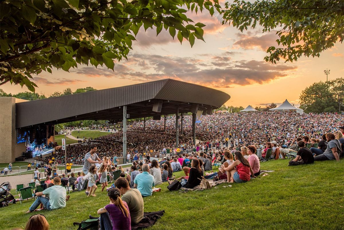 Outdoor concert at sunset with a large crowd seated on the grassy hillside, stage with performers under a covered structure, and tents in the background.
