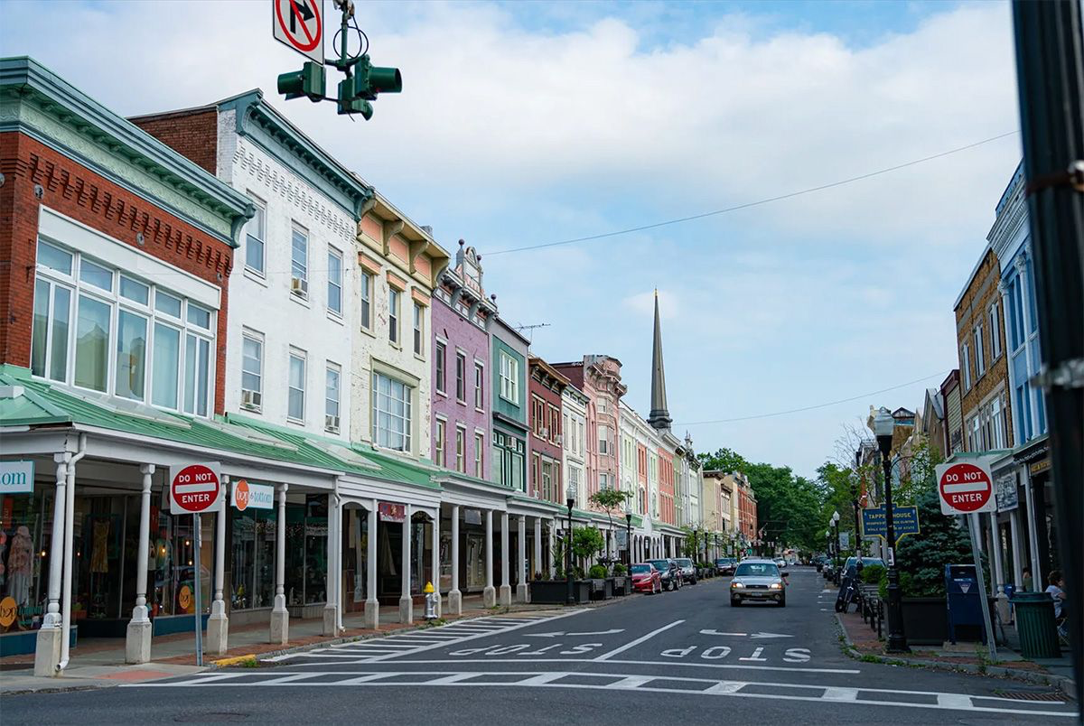 Colorful row of historic storefronts and residential buildings on a small town street with a church steeple in the background, under a partly cloudy sky.
